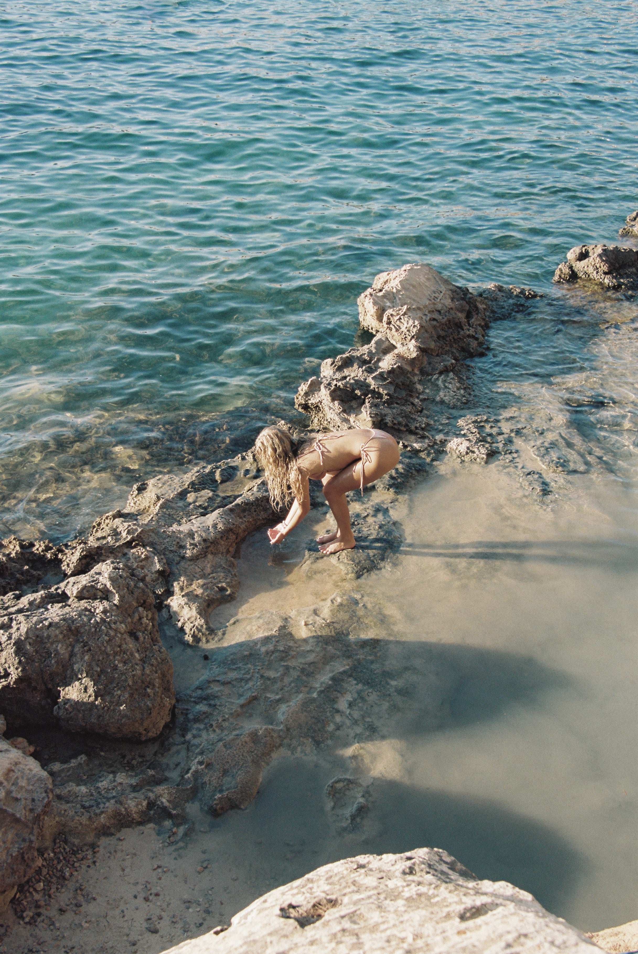 woman in nude string bikini crouching by tide pool on rocky shoreline